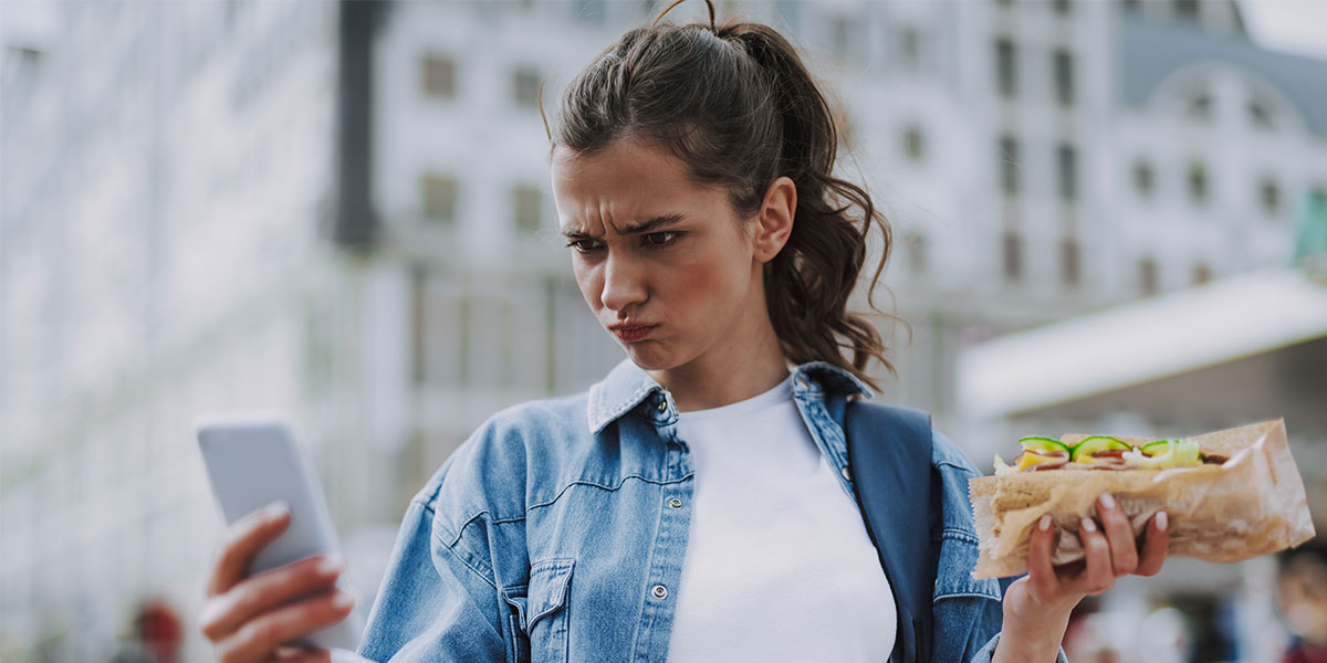 Female holding a sandwich, frowning, while holding her mobile phone